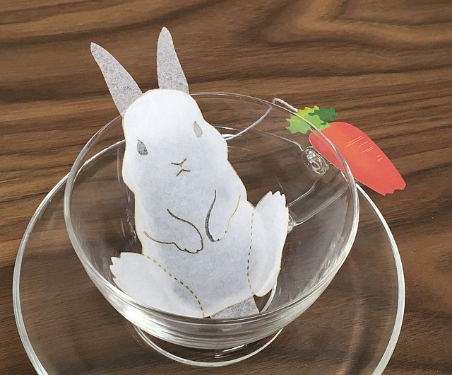 a clear teacup with a tea packet in the shape of a rabbit inside of it, the end of the tea packet has a paper carrot